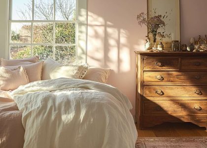 Modern French country bedroom with sunlight through large window, pink-hued walls, weathered oak bed frame with fluffy white duvet and pastel pillows, antique reclaimed wood dresser with trinkets, and plush area rug for a tranquil relaxation haven.