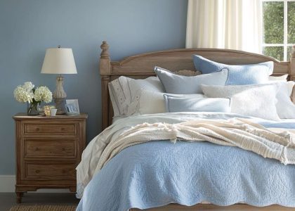 Modern French country bedroom with soft blue walls, sturdy wooden bed, layered cream and blue bedding, rustic vintage bedside table, woven jute rug, and sunlight streaming through billowy curtains.