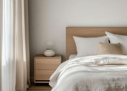 Minimalist bedroom with soft beige walls, wooden bed frame, lightweight white duvet, single decorative pillow, natural wood bedside table, ceramic lamp, and sheer curtains filtering morning light.