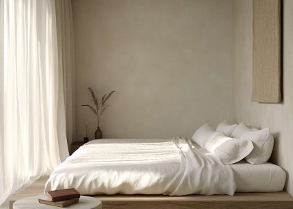 Minimalist bedroom with a light wood bed against a beige wall, featuring white linens and earth-toned throw pillows, a natural stone side table with a vintage book and lamp, and sheer curtains allowing natural light to create a serene atmosphere.